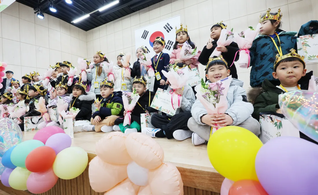 First-year students sit on a stage at an elementary school entrance ceremony in Korea, wearing paper crowns and holding flower bouquets. Photo: Kyunghyang Shinmun